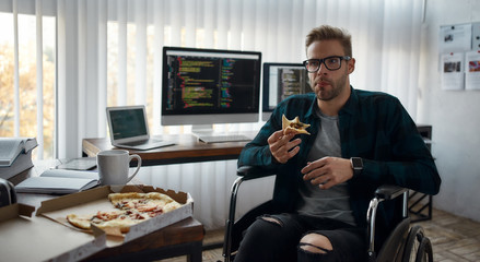 So delicious. Portrait of young hungry male web developer in a wheelchair eating pizza while sitting at his workplace in the office. Multiple computer screens with program code