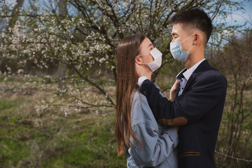 Asian guy with a European girl in medical masks on the background of flowering trees hug and kiss