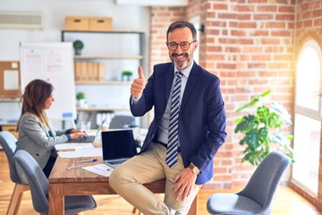 Middle age handsome businessman wearing glasses sitting on desk at the office doing happy thumbs up...