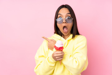 Young brunette girl holding a cornet ice cream over isolated pink background surprised and pointing side