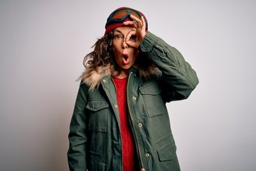 Middle age skier woman wearing snow sportswear and ski goggles over white background doing ok gesture shocked with surprised face, eye looking through fingers. Unbelieving expression.