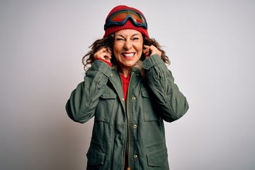 Middle age skier woman wearing snow sportswear and ski goggles over white background covering ears with fingers with annoyed expression for the noise of loud music. Deaf concept.
