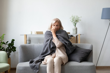 Unhealthy middle aged woman covered in blanket sitting on sofa in living room. Sick older lady caught cold, suffering from fever or allergic infection reaction, wiping runny nose with handkerchief.