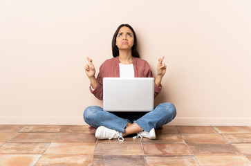 Young mixed race woman with a laptop sitting on the floor with fingers crossing and wishing the best