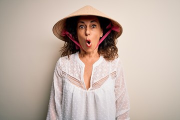 Middle age brunette woman wearing asian traditional conical hat over white background afraid and shocked with surprise expression, fear and excited face.