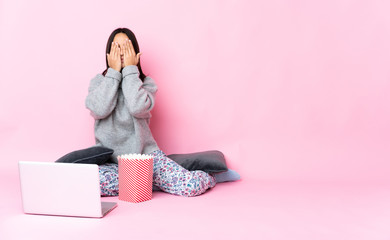 Young mixed race woman eating popcorn while watching a movie on the laptop covering eyes by hands