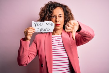 Middle age woman holding paper with you are fired message over isolated pink background with angry...