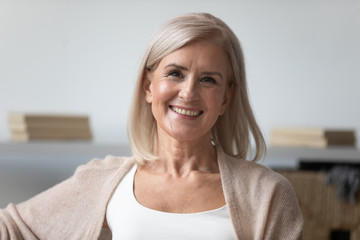 Head shot close up portrait of pleasant smiling mature woman. Happy healthy middle aged lady relaxing alone at home, looking at camera, posing for photo. Positive satisfied retirement concept.