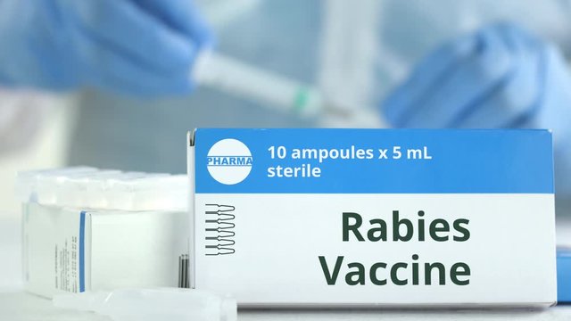 Box with rabies vaccine on the table against blurred lab assistant or doctor. Fictional phaceutical logo