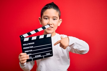 Young little boy kid filming video holding cinema director clapboard over isolated red background...