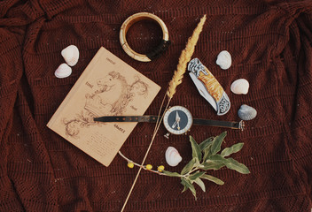 Notepad, compass, knife, olive branch, spikelet, bone bracelet and shells on a brown bedspread in the sand.