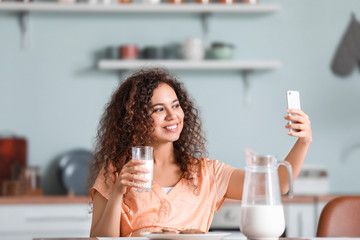 Young African-American woman with milk taking selfie in kitchen