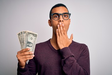 Handsome african american man holding bunch of dollars banknotes over white background cover mouth with hand shocked with shame for mistake, expression of fear, scared in silence, secret concept