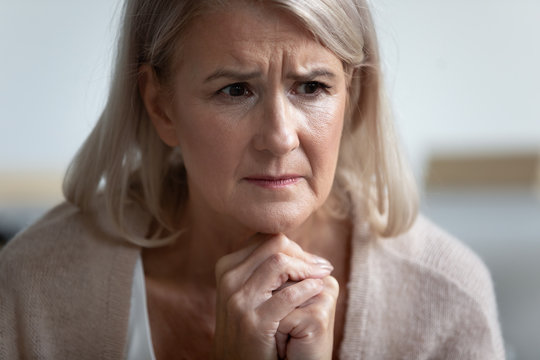 Frowning upset mature woman put head on folded hands, looking in distance. Lost in thoughts upset woman sitting alone, head shot close up portrait. Mental disorder or dementia, health problem concept.