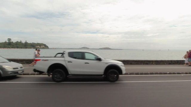 Driving on a road next to Ocean Bay in Auckland, New Zealand, passing by cars parked next to the road and people walking by.