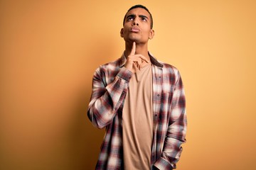 Young handsome african american man wearing casual shirt standing over yellow background Thinking concentrated about doubt with finger on chin and looking up wondering