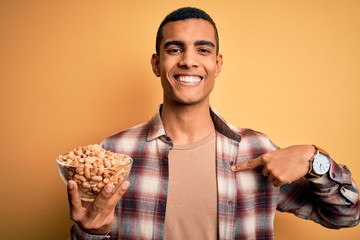 Handsome african american man holding bowl with heathy peanuts over yellow background with surprise face pointing finger to himself