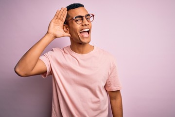 Handsome african american man wearing casual t-shirt and glasses over pink background smiling with hand over ear listening an hearing to rumor or gossip. Deafness concept.