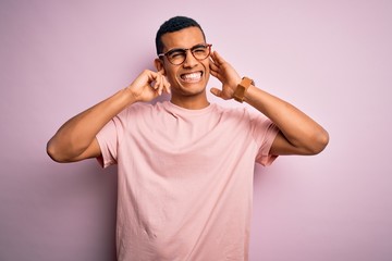 Handsome african american man wearing casual t-shirt and glasses over pink background covering ears with fingers with annoyed expression for the noise of loud music. Deaf concept.