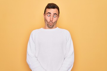 Young handsome man with blue eyes wearing casual sweater standing over yellow background making fish face with lips, crazy and comical gesture. Funny expression.