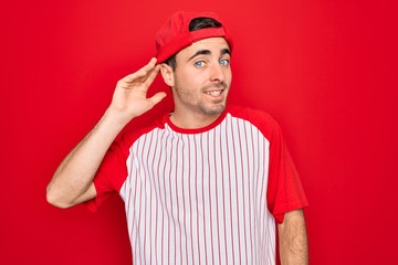 Young handsome sporty man with blue eyes wearing striped baseball t-shirt and cap smiling with hand over ear listening an hearing to rumor or gossip. Deafness concept.