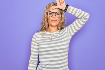 Beautiful blonde woman wearing casual striped t-shirt and glasses over purple background making fun of people with fingers on forehead doing loser gesture mocking and insulting.