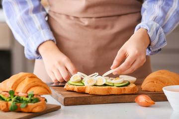 Obraz na płótnie Canvas Woman preparing tasty croissant sandwich in kitchen, closeup
