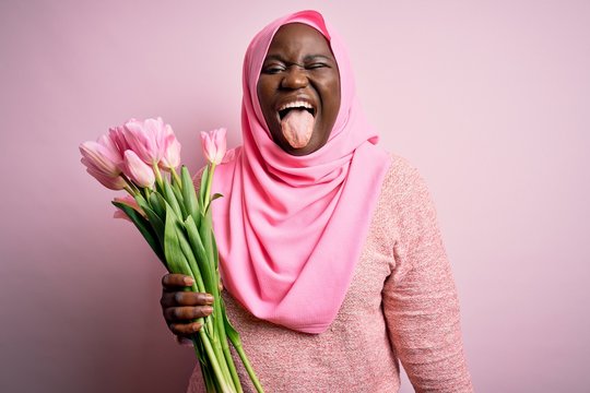 Young african american plus size woman wearing muslim hijab holding bouquet of pink tulips sticking tongue out happy with funny expression. Emotion concept.