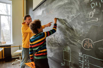 The great aim of education is not knowledge but action. Young male science teacher explaining main rules and formulas in physics and mechanics. A boy is drawing on the blackboard - Powered by Adobe
