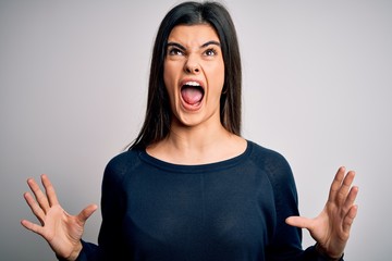 Young beautiful brunette woman wearing casual sweater standing over white background crazy and mad shouting and yelling with aggressive expression and arms raised. Frustration concept.