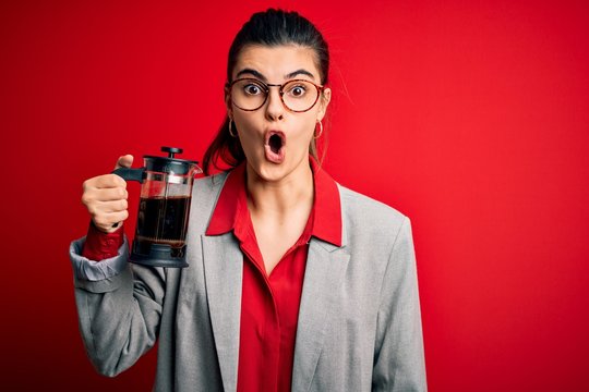 Young beautiful brunette woman doing coffe holding french coffeemaker over red background scared in shock with a surprise face, afraid and excited with fear expression