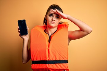 Young beautiful brunette woman wearing safe lifejacket showing smartphone stressed with hand on...