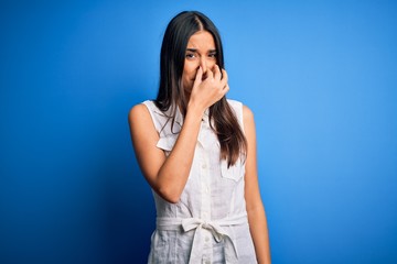 Young beautiful brunette woman wearing casual dress over isolated blue background smelling...