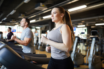 Waist up portrait of smiling young woman running on treadmill while enjoying cardio workout in modern gym, copy space