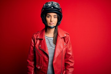 Young beautiful brunette motorcyclist woman wearing motorcycle helmet and red jacket with serious expression on face. Simple and natural looking at the camera.