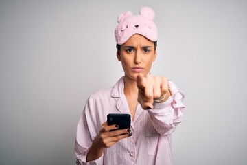 Young beautiful woman wearing pajama and sleep mask having conversation using smartphone pointing with finger to the camera and to you, hand sign, positive and confident gesture from the front