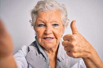 Senior beautiful grey-haired woman making selfie by camera over isolated white background happy with big smile doing ok sign, thumb up with fingers, excellent sign