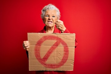 Senior beautiful grey-haired woman holding banner with prohibited signal over red background...