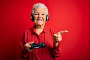 Senior beautiful grey-haired gamer woman playing video game using joystick and headphones very happy pointing with hand and finger to the side © Krakenimages.com