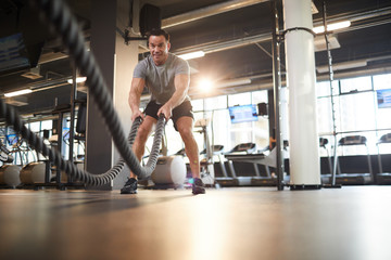 Fototapeta na wymiar Full length wide angle portrait of mature muscular man doing battle rope exercises during cross workout in modern gym lit by sunlight, copy space