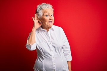 Senior beautiful woman wearing elegant shirt standing over isolated red background smiling with hand over ear listening an hearing to rumor or gossip. Deafness concept.