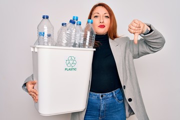 Young beautiful redhead woman recycling holding trash can with plastic bottles to recycle with angry face, negative sign showing dislike with thumbs down, rejection concept