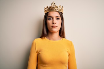 Young beautiful brunette woman wearing golden queen crown over white background with serious expression on face. Simple and natural looking at the camera.