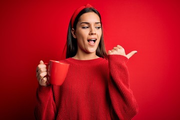 Young beautiful brunette woman drinking mug of coffe over isolated red background pointing and...