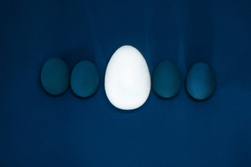 blue eggs stand in a row on a plain blue background, in the center is a large white egg, makings of a leader, Easter concept. image is dark