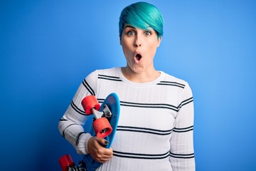 Young skater woman with blue fashion hair holding skateboard over blue isolated background scared in shock with a surprise face, afraid and excited with fear expression