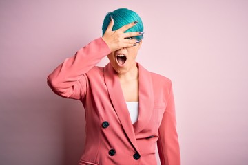 Young beautiful businesswoman with blue fashion hair wearing jacket over pink background peeking in shock covering face and eyes with hand, looking through fingers with embarrassed expression.