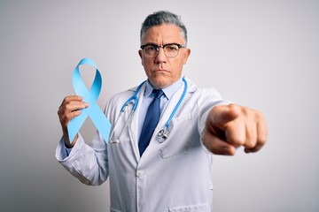 Middle age handsome grey-haired doctor man wearing coat holding blue cancer ribbon pointing with finger to the camera and to you, hand sign, positive and confident gesture from the front