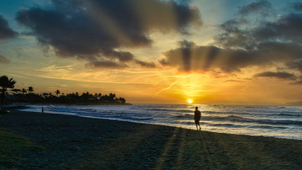 Fototapeta premium Silhouette of a man walking on a beach at sunrise with suns rays across the sky
