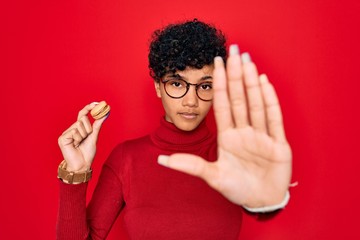 Young beautiful african american afro woman holding chocolate french dessert macaron with open hand doing stop sign with serious and confident expression, defense gesture